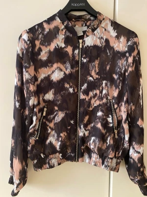 Silky jacka - Snygg bomberjacka från HM med abstrakt mönster i svart, beige och vitt. Jackan har dragkedja framtill, två fickor med guldfärgade dragkedjor och resår i midja och ärmslut. Lätt material, perfekt för lager på lager och trendig look.