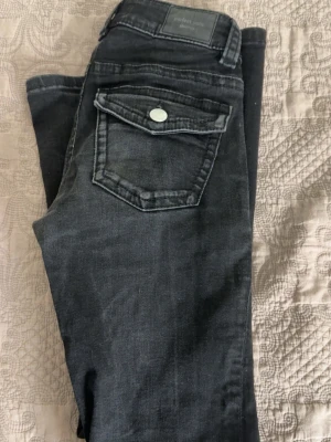 Svarta jeans med ficklock - Snygga svarta jeans med normal midja och raka ben. Jeansen har en stor ficka med lock och knapp på baksidan samt klassiska detaljer framtill. Materialet är mjukt denim som ger en skön känsla och passar till många olika stilar.