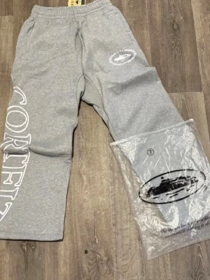 Grå mjukisbyxor från Corteiz - Snygga grå mjukisbyxor från Corteiz med stort vitt tryck längs benet och logga på höften. Byxorna har resår i midjan och sidofickor. Tillverkade i mjukt bomullsmaterial som är perfekt för chill dagar. Passar dig som gillar streetwear och vill ha något bekvämt men ändå stilrent.