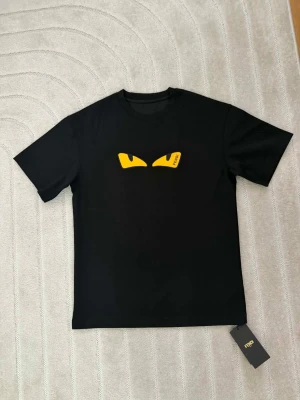 Svart Fendi t-shirt med gula ögon - Snygg svart t-shirt från Fendi med ikoniska gula ögon på bröstet och diskret Fendi-logga i trycket. Klassisk passform och rund halsringning. Perfekt för dig som vill sticka ut med en exklusiv streetwear-look.