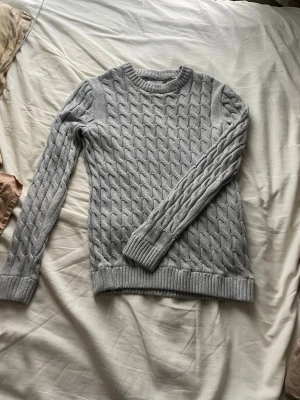 Ljusgrå kabelstickad tröja Only & Sons - Snygg ljusgrå kabelstickad tröja från Only & Sons. Tröjan har rund halsringning, ribbade muddar och ett klassiskt flätmönster över hela plagget. Perfekt för dig som gillar en stilren och enkel look med lite extra struktur.