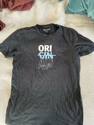 Svart t-shirt Jack & Jones Originals - Svart t-shirt från Jack & Jones Originals med tryck i vitt och blått på bröstet. Klassisk rund hals och korta ärmar. Tillverkad i mjuk bomull för en skön känsla. Perfekt för en avslappnad och stilren look. Lätt använd och inga defekter. 