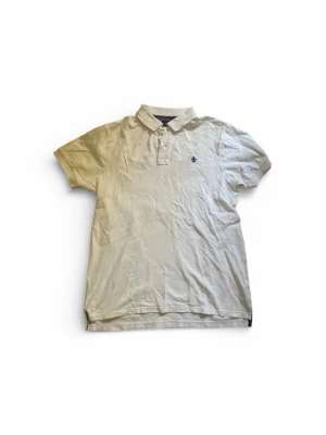 Vit pikétröja Morris  - Vit pikétröja från Morris Stockholm i storlek M. Klassisk design med krage, korta ärmar och två knappar framtill. Broderad mörkblå logga på bröstet. Materialet är mjuk bomull och insidan av kragen har lila tyg med vita prickar för en extra detalj.