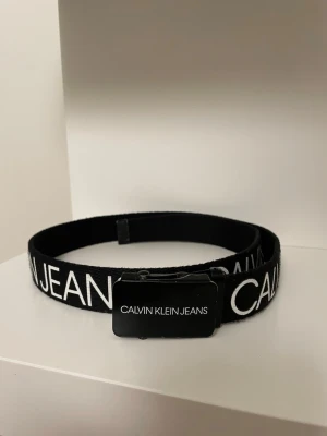 Svart bälte från Calvin Klein Jeans - Snyggt svart bälte från Calvin Klein Jeans med vit logotyptext längs hela bandet. Bältet har ett modernt spänne i svart metall med märkets namn tryckt på. Perfekt accessoar för att lyfta din outfit.