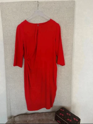 Röd klänning med trekvartsärm - Snygg röd klänning med trekvartsärm och draperad detalj i sidan. Klänningen har rund halsringning och en enkel, elegant siluett. Perfekt för dig som vill sticka ut med färg. Materialet känns mjukt och följsamt.