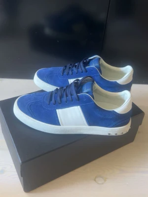 Blåa sneakers från Valentino - Snygga blåa sneakers från Valentino med vita detaljer och svart sula. Ovandelen är i mocka och läder, med svarta skosnören och en vit panel på sidan. Klassisk låg modell som passar till många olika stilar. PASSAR 42 och 43!