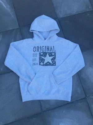 Original tryck hoodie grå - Tjeenaaaaaa!!! Äntligen sommarlov och jag säljer nu en Ljusgrå hoodie från Original Fit med stor huva och magficka. Tryck på bröstet med texten 'ORIGINAL FIT EST. 2024' och en stor stjärna omgiven av mindre stjärnor. Mjuk bomullsblandning och avslappnad passform. Perfekt hoodie för alla tillställningar och är bara väldigt snygg och passar till allt!! Storlek S skick 9,5/10 se bild en minimal fläck på ena armslutet snören saknas tyvärr 😁hör av dig vid mer frågor eller funderingar🤩mvh Ga🕺