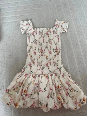 Blommig kortklänning med volang - Söt kortklänning med blommigt mönster i rosa, grönt och gult på vit botten. Klänningen har puffiga korta ärmar, smockad överdel och volangig kjol. Perfekt för sommarens alla tillfällen!