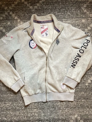 Grå zip-hoodie från U.S. Polo Assn. - Grå zip up tröja med dragkedja från U.S. Polo Assn. Snygga broderade och tryckta detaljer på bröstet, ärmen och axeln. Ribbstickade muddar och krage, samt två fickor framtill. Perfekt för en sportig och avslappnad stil. Storlek XL och bra skick