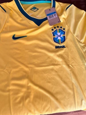 Brasilien Nike fotbollströja gul - Gul Brasilien fotbollströja från Nike med gröna detaljer och broderat CBF-emblem på bröstet. Tröjan har rund hals med grönt kantband och meshpaneler på sidorna för extra ventilation. Perfekt för dig som älskar fotboll och vill stötta Brasilien.