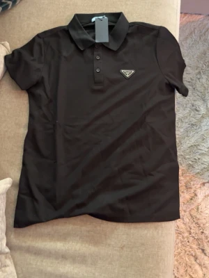 Svart pikétröja från Prada - Säljer en svart pikétröja från Prada med klassisk krage och knappslå framtill. Tröjan har korta ärmar och en diskret logga på bröstet. Tillverkad i mjuk bomull som känns skön mot huden. Perfekt för en clean och stilren look. Storlek är medium men den är absolut inte medium, den är typ S eller XS. Passade inte mig så väljer nu att svälja 👍