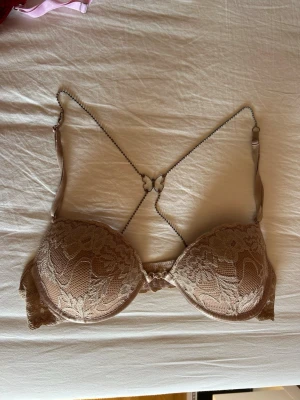 Beige bh med spetsdetaljer - Snygg beige push-up bh med vadderade kupor och vacker spets framtill. Axelbanden är justerbara och korsas i ryggen med en dekorativ fjärilsdetalj. Perfekt för dig som vill ha både stöd och en feminin look.