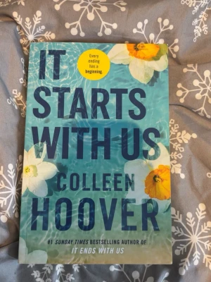 It Starts With Us av Colleen Hoover - En skönlitterär bok med blått omslag och vita samt gula blommor. Titeln 'It Starts With Us' är tryckt i stora bokstäver och författaren är Colleen Hoover. Boken är på engelska och har ett mjukt omslag.