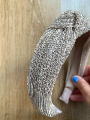 Beige metallic diadem med knut - Snyggt diadem i beige med metallicskimmer och en trendig knut på toppen. Tillverkat i ett glansigt tyg som ger en lyxig känsla och passar perfekt för att lyfta vilken outfit som helst. Bekväm insida med mjuk vaddering.