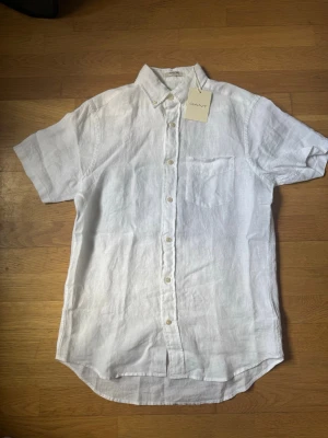 Vit linneskjorta från Gant - Klassisk vit skjorta från Gant i svalt linne. Skjortan har korta ärmar, bröstficka och knappar framtill. Perfekt för varma dagar och enkel att styla med allt. Regular fit och tidlös design.
