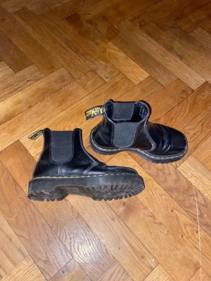 Svarta Chelsea boots Dr. Martens - Säljer ett par klassiska svarta Chelsea boots från Dr. Martens. Skorna har elastiska paneler på sidorna, grov sula och den ikoniska gula sömmen runt sulan. Tillverkade i slitstarkt läder och har dragflik med logga bak.