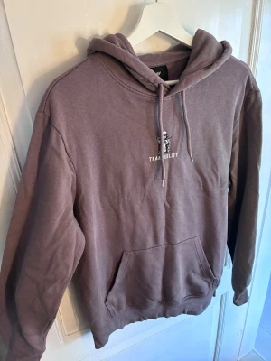 Ljusbrun hoodie från H&M med tryck - Säljer en ljusbrun hoodie från H&M med ett litet tryck av en ängel och texten 'TRANQUILITY' på bröstet. Hoodien har huva med dragsko och en stor magficka. Regular fit och mjukt material, perfekt för chill dagar.