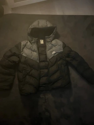 Svart Nike pufferjacka med huva barnstorlek XL - Säljer en svart pufferjacka från Nike med Therma-Fit-teknologi. Jackan har en stor huva, dragkedja framtill och gråa paneler över axlarna. Nike-logga på bröstet. Perfekt för kalla dagar och riktigt skön att bära.