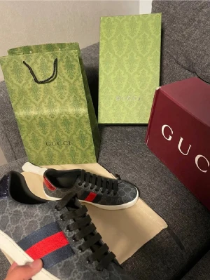Gucci sneakers med monogram och röd/blå rand - Snygga Gucci sneakers i svart med klassiskt GG-monogram och ikoniska röd/blå ränder på sidorna. Skorna har svarta skosnören, vit sula och är tillverkade i canvas med läderdetaljer. Perfekta för dig som vill sticka ut med lyxig streetstyle.
