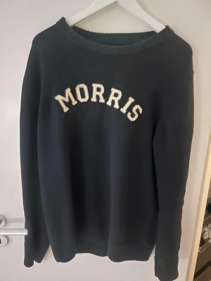 Mörkblå sweatshirt från Morris - Mörkblå sweatshirt från Morris med vit broderad logga på bröstet och en patch med brittiska flaggan på ärmen. Klassisk rund halsringning och mjukt material som passar perfekt till jeans eller chinos.