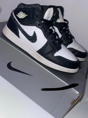Air Jordan 1 Mid svart/vit sneakers - Snygga Air Jordan 1 Mid sneakers i svart och vitt läder med klassisk Air Jordan-logga på sidan och mönstrad Swoosh. Skorna har svart/vita paneler och coola detaljer runt kragen. Perfekta för dig som gillar streetwear och vill sticka ut.