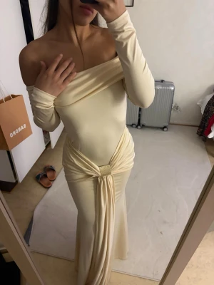Lång offshoulder klänning i beige - Elegant långklänning i beige med offshoulder-design och långa ärmar. Klänningen har en snygg drapering och knytdetalj i midjan som ger en unik look. Tillverkad i ett mjukt, stretchigt material som sitter slimmat mot kroppen.