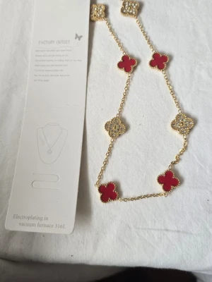 Guldhalsband med röda och pärlformade detaljer - Snyggt halsband med guldfärgad kedja och fyrklöverformade hängen i rött och gnistrande pärlformade detaljer. Kedjan är tunn och elegant, och hänget växlar mellan röda emaljdelar och glittrande stenar för en lyxig look.