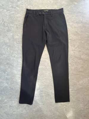 Kostymbyxor Tailored Originals - Snygga marinblå kostymbyxor från Tailored Originals med smal passform och klassisk design. Byxorna har två sidofickor, bälteshällor och knappgylf. Perfekta för en clean och stilren look.