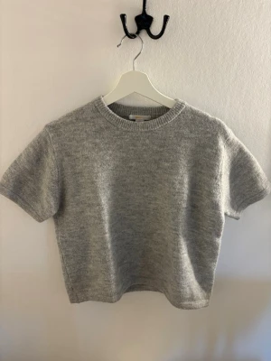 Grå stickad t-shirt från Gina Tricot - Mysig grå stickad t-shirt från Gina Tricot med korta ärmar och rund halsringning. Tröjan har en enkel och stilren design i mjukt material, perfekt för lager på lager eller som den är. Perfekt att ha ett spets linne under!🩷 Aldrig använd