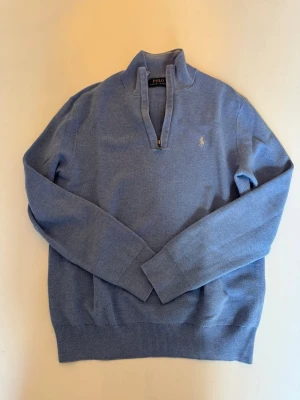 Blå halvziptröja Polo Ralph Lauren - Ljusblå stickad tröja från Polo Ralph Lauren med halvzip och ståkrage. Tröjan har lång ärm, ribbade muddar och den klassiska broderade loggan på bröstet. Perfekt för en clean och avslappnad stil. Mycket fint skick, endast använd ett fåtal gånger. 