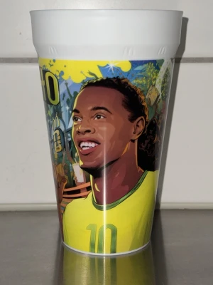 Plastmugg med Ronaldinho-motiv - Färgglad plastmugg med tecknad bild av fotbollsspelaren Ronaldinho i brasiliansk landslagströja. Muggen har tryck med nummer 10 och texten 'RONALDINHO Perfekt för fotbollsälskare eller som samlarobjekt.