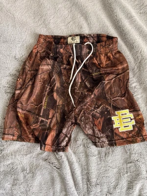 Camo shorts från EEC med snörning - Säljer ett par camouflagemönstrade shorts från EEC, tillverkade i New York. Shortsen har elastisk midja med vit snörning och meshmaterial som andas. Färgskalan går i brunt och grönt med gul logga på benet. Perfekta för sommaren!