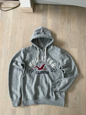 Grå hoodie från Hollister - Snygg grå hoodie från Hollister med stor broderad logga och texten 'California' på bröstet. Hoodien har dragsko i huvan och ribbade muddar vid ärmar och nederkant. Tillverkad i mjukt sweatshirtmaterial, perfekt för en avslappnad stil. Finns ett hål som knappt syns (se bild).