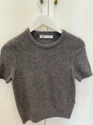 Grå stickad topp från Zara - Mysig grå stickad topp från Zara med korta ärmar och rund halsringning. Perfekt att styla med jeans eller kjol för en chill och trendig look. Materialet känns mjukt och skönt mot huden.