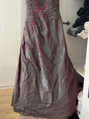 Lång vinröd balklänning med broderi - Elegant långklänning i vinröd och silverton med vackra broderade blommor och paljettdetaljer upptill. Klänningen har en åtsittande överdel med rynk och vid kjol med flera lager tyg, inklusive svart tyll underkjol. Perfekt för bal eller festliga tillfällen.