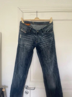 Blåa jeans med broderade bakfickor - Säljer ett par blå jeans med slitningar och coola broderade detaljer på bakfickorna. Jeansen har klassisk femficksmodell, raka ben och tydliga whiskers-effekter framtill. Perfekta för dig som gillar en avslappnad och trendig stil.