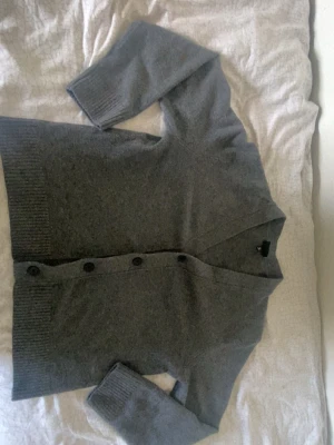 Grå cardigan - Mysig grå kofta med v-ringning och stora svarta knappar framtill. Koftan har ribbstickade muddar vid ärmslut och nederkant, samt en avslappnad passform. Perfekt att slänga över en t-shirt för en chill look.