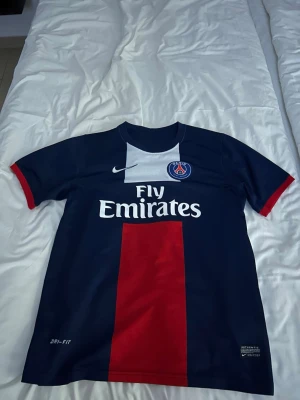 PSG matchtröja Ibrahimović #10 Nike - Säljer en mörkblå PSG matchtröja från Nike med röd mittpanel och vita detaljer. Tröjan har trycket 'Fly Emirates' på bröstet, PSG-logga och Nike-märke. På ryggen står det Ibrahimović och nummer 10. Tillverkad i Dri-FIT material för bästa komfort.