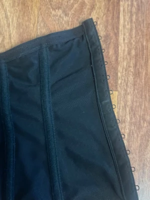 Svart korsett med spetsdetaljer - Snygg svart korsett med dekorativa guldfärgade spetsbroderier på framsidan. Modellen har markerade sömmar och hakfästen längs sidan för justerbar passform. Materialet är en mix av tyg och mesh med förstärkta partier för extra stöd.