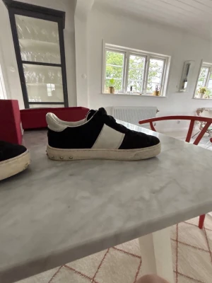 Svarta och vita sneakers med mockadetaljer - Snygga valentino föycrews i svart mocka med vita läderdetaljer och vit sula. Klassisk låg modell med svarta skosnören och diskreta nitar på sulan. Perfekta för en stilren och avslappnad look.