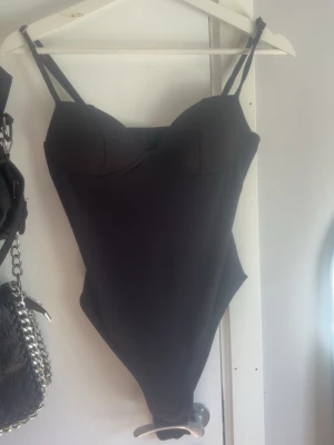 Svart body med bygel och smala band - Säljer en svart body med bygelkupor och smala axelband. Bodyn har en enkel och stilren design med lätt vadderade kupor och en liten rosett framtill. Materialet känns stretchigt och följsamt, perfekt för att skapa en snygg siluett.