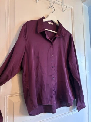 Lila skjorta från JDY med knappar - Snygg lila skjorta från JDY med klassisk krage och knäppning framtill. Skjortan har lång ärm och är tillverkad i ett glansigt, mjukt material som ger en elegant känsla. Perfekt att styla till jeans eller kjol för en trendig look.