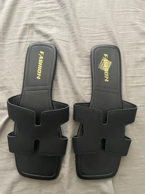 Svarta slip-in sandaler med fyrkantig tå - Stilrena svarta slip-in sandaler från Fashion med fyrkantig tå och breda remmar i konstläder. Snyggt H-format mönster på ovandelen och bekväm platt sula. Perfekta för en trendig och avslappnad look. Storlek 40 men passar 39 också. Aldrig använda ute. Endast testade hemma. 