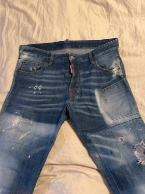 Blå slitna jeans från Dsquared2 - Säljer ett par blå jeans från Dsquared2 med coola slitningar och tvättade detaljer. Jeansen har fem fickor, knappgylf och klassisk passform. Tillverkade i Italien av denim med snygga slitningar och patchade partier för en trendig look.
