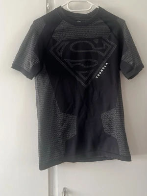  Superman compression T-shirt  YoungLA -  svart t-shirt från YoungLA med stort Superman-tryck på bröstet och gråa mönstrade paneler på ärmar och sidor. T-shirten har rund hals och korta ärmar, perfekt för dig som gillar superhjältar och streetwear.
