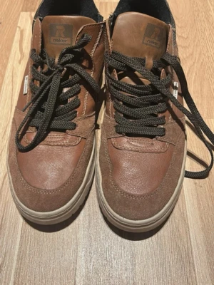 Bruna sneakers från Rieker - Snygga bruna sneakers från Rieker med både mocka- och läderdetaljer. Skorna har svarta snören, dragkedja på sidan och en beige sula. Perfekta för dig som gillar en klassisk men ändå modern look.