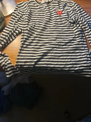 Randig långärmad  - Säljer en klassisk långärmad topp frånmed svartvita ränder och det ikoniska röda hjärtat på bröstet. Toppen har en avslappnad passform och är tillverkad i mjuk bomull, perfekt för en stilren vardagslook. En liten orange fläck som syns i speciellt ljus men inga andra defekter