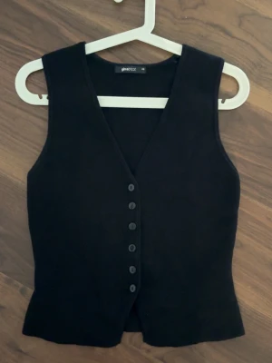 Svart väst från Gina Tricot - Stilren svart väst från Gina Tricot med djup V-ringning och sex knappar framtill. Västen har en figurnära passform och är tillverkad i ett mjukt tyg som ger en klassisk look. Endast använd en gång 🩷