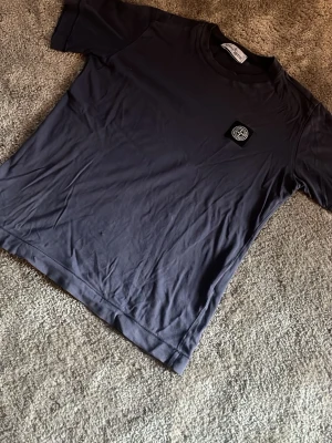 Lila grå  t-shirt från Stone Island Junior - Snygg lila grå t-shirt från Stone Island Junior med klassisk rund hals och ikonisk patch på bröstet. Tillverkad i mjuk bomull som känns skön mot huden. Perfekt för dig som gillar stilrena och trendiga plagg med streetwear-vibe.