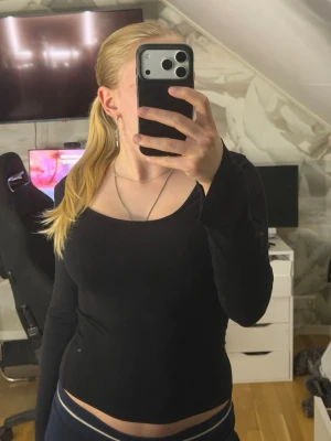 Svart långärmad topp med rundringning - En enkel och stilren svart långärmad topp med djup rundringning. Toppen har en tight passform och är tillverkad i ett mjukt stretchigt material som sitter snyggt på kroppen. Perfekt att matcha med jeans eller kjol för en clean look.